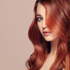 Ein Porträt einer Frau mit voluminösen, dunkelroten Haaren, die ihr Gesicht umspielen. Ihre blauen Augen blicken nachdenklich in die Ferne.