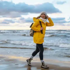 Uśmiechnięta kobieta w żółtej kurtce spaceruje po plaży. Wiatr rozwiewa jej włosy. To piękny jesienny dzień nad morzem.