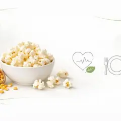 Miseczka popcornu, obok drewniana łyżka z ziarnami kukurydzy. Ikony serca z liściem i talerza z sztućcami sugerują zdrowe przekąski i ich popcorn kcal.