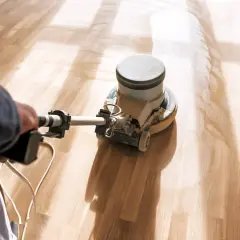 En person slipar parkettgolv med en maskin. Solstrålar lyser upp det nyslipade träet.