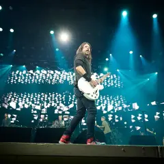 Dave Grohl z Foo Fighters gra na gitarze podczas koncertu, oświetlony niebieskimi światłami. Chce pokazać, jak grać.