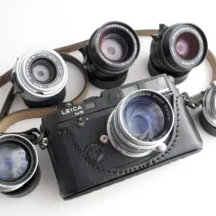 A Leica M6 camera with a leather case is surrounded by several best M mount lenses, showcasing a collection of classic photographic gear.