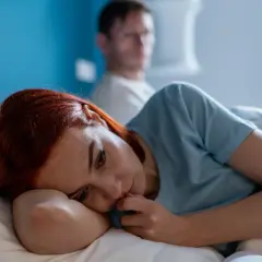Smutna kobieta leży w łóżku, czując się jak ofiara w związku. Jej partner siedzi za nią, odwrócony plecami.