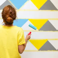 Frau mit rotem Dutt streicht mit Roller geometrische Muster an Wand. Ideen für Wand streichen mit Muster abkleben.