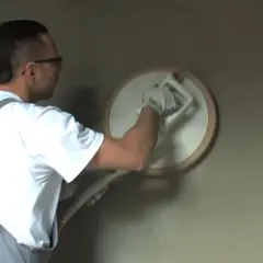 Pracownik w ogrodniczkach wykonuje zacieranie tynku cementowo wapiennego za pomocą kielni.