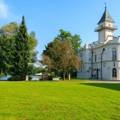 Biały zamek z wieżą, otoczony zielonym parkiem i jeziorem. Urokliwe zamki na Mazowszu zachwycają.