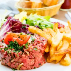 Un délicieux tartare de boeuf, garni d'une tranche de tomate et de persil, servi avec des frites croustillantes et une salade fraîche.