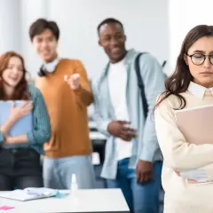 Smutna dziewczyna z okularami trzymająca teczkę, podczas gdy grupa ludzi śmieje się i wskazuje na nią. To przykład dyskryminacji w pracy.