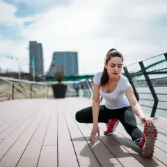 Kobieta wykonuje dynamiczny stretching na drewnianym tarasie nad rzeką, w tle miejski krajobraz z mostem.