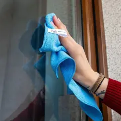 Dłoń z niebieską ściereczką z mikrofibry czyści okno, by lśniło. Poznaj sekret, czym myć okna, żeby się błyszczały bez smug.