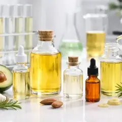 Butelki z olejkami, w tym potencjalnie hydrogenated vegetable oil, obok awokado, kokosa i migdałów. W tle probówki i mikroskop.