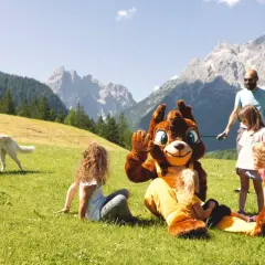Famiglia al family resort Rainer Sesto, con mascotte e cane su prato verde, montagne sullo sfondo.
