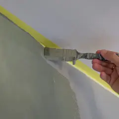 En hand målar en vägg med en pensel, noggrant längs maskeringstejpen. Snart är rummet färdigmålat!