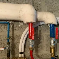 Tuyauterie gainée pour le calorifugeage, avec vannes rouges et bleues sur fond de mur en parpaings.