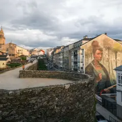Mural de una mujer en la muralla de la fortaleza Lugo, con la catedral al fondo y un cielo nublado.