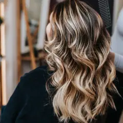 Mujer con cabello castaño y mechas rubias ondulado, visto desde atrás en un salón de belleza.