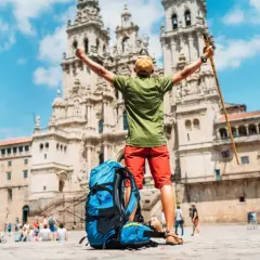 Pielgrzym z plecakiem i laską triumfalnie wznosi ręce przed katedrą w Santiago de Compostela, kończąc swój szlak.