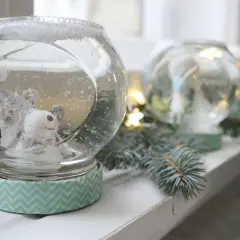 Urocza kula śnieżna diy z misiem w środku, otoczona gałązkami choinki i światełkami.