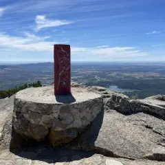 Pilar rojo con grafitis en la cima de una montaña, con un paisaje de valle y lago al fondo. La **cabeza lijar** del pilar parece desgastada por el tiempo.