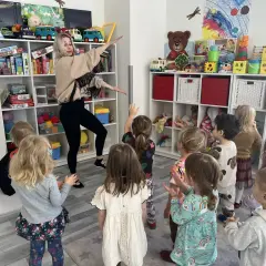 Nauczycielka z przedszkola prowadzi zajęcia rytmiczne dla grupy dzieci, które patrzą na nią z zaciekawieniem.