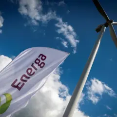 Flaga z logo Energa powiewa na tle błękitnego nieba z chmurami, obok wiatraka. Akcje Energa rosną.