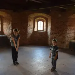 Dzieci zwiedzają wnętrze z ceglanymi ścianami i zabytkowymi skrzyniami w zamku krzyżackim w Bytowie.