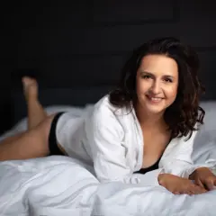 Uśmiechnięta kobieta w białej koszuli leży na łóżku. Świetne pomysły na sesję w domu.