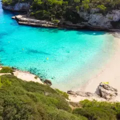 Traumhafte **Buchten Mallorcas** mit türkisfarbenem Wasser, weißen Sandstränden und grünen Pinienwäldern. Ein Boot ankert im Meer, ein Rettungsschwimmerturm steht am Strand.