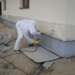 Pracownik w kombinezonie wykonuje malowanie elewacji po ociepleniu styropianem, zabezpieczając dolną część budynku folią.