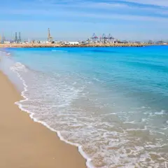 Olas suaves acarician la arena dorada en las **playas de Valencia**, con el puerto industrial de fondo bajo un cielo azul.