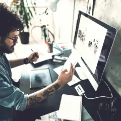 Ein Mann mit lockigen Haaren und Tattoos arbeitet an seinem Computer. Er ist im Bereich IU Kommunikationsdesign tätig und entwirft visuelle Konzepte.