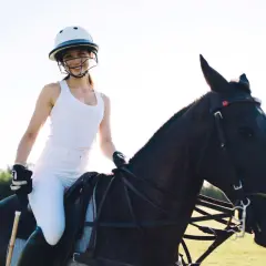 Uśmiechnięta kobieta w kasku i białym stroju do jazdy konnej w terenie, siedząca na ciemnym koniu.