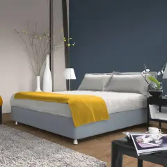 Ein Schlafzimmer mit einem Bett, das mit einer gelben Decke und Kissen in verschiedenen **blau nuancen** dekoriert ist. Ein gelber Sessel und ein Teppich ergänzen den Raum.