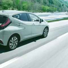 Jasnoszary Toyota Aqua jedzie po krętej drodze wśród zielonych wzgórz.