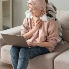 Kobieta z dzieckiem na kolanach pracuje przy laptopie. Dziecko ją przytula, szukając wsparcia, jak u psychoonkologa.