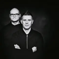 Dwóch mężczyzn w czarnych bluzach na ciemnym tle. Jeden z nich ma okulary, drugi irokeza. Wyglądają jakby właśnie wylądowali po locie dzika.