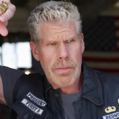 Ron Perlman w skórzanej kamizelce z naszywkami, z tatuażem na ramieniu i pierścieniami na palcach, na tle amerykańskiej flagi.