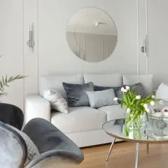 Przytulny salon z jasnoszarymi, **białymi ścianami**, wygodną kanapą, welurowymi fotelami i okrągłym lustrem.
