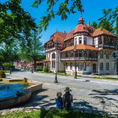 Dwie osoby siedzą przy fontannie, podziwiając architekturę luksusowego sanatorium w otoczeniu zieleni.