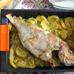 Pargo entero al horno con patatas y cebolla en bandeja negra con asas naranjas.