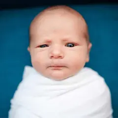 Un neonato senza capelli davanti, avvolto in una coperta bianca, guarda con occhi curiosi.