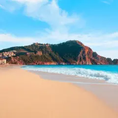 Plaża Kleopatry w Alanyi, z piaszczystym brzegiem, turkusowym morzem i skalistym wzgórzem z zamkiem.