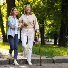 Młoda kobieta pomaga starszej pani, która wspiera się na czwórnodze, spacerując po parku.