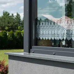 Szary parapet okienny z kamienia, z ozdobną koronkową firanką wewnątrz. Za oknem zielony ogród.