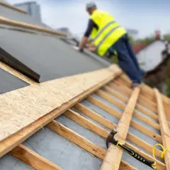 Pracownik na dachu układa blachę z membraną. Widoczny młotek i gwoździe.