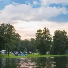 Malowniczy pole campingowe nad jeziorem, z namiotami, samochodami i ludźmi odpoczywającymi wśród drzew.