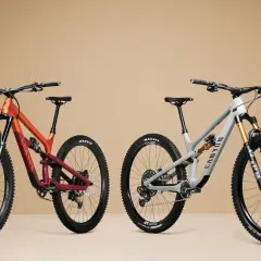 Two full-suspension mountain bikes, one orange and burgundy, the other grey, are shown side-by-side, highlighting the 27.5 vs 29 wheel size debate.
