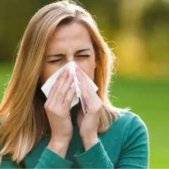 Mujer rubia con pañuelo en la nariz, sufriendo síntomas de la rinitis alérgica en un día soleado.