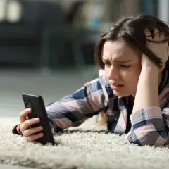 Zmartwiona dziewczyna leży na dywanie, próbując włączyć telefon, który nie chce się włączyć.