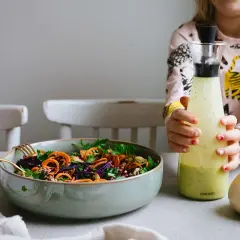 En färgstark sallad med morötter och rödkål i en skål, bredvid två flaskor med dressing till sallad. Ett barn håller i den ena flaskan.